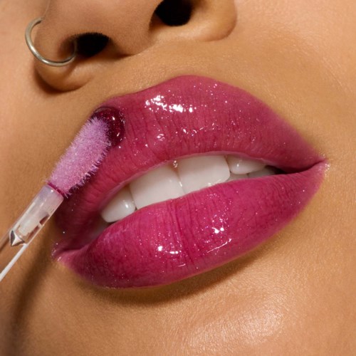 Een persoon brengt roze lipgloss aan met een kwast, toont volle lippen en neuspiercing.