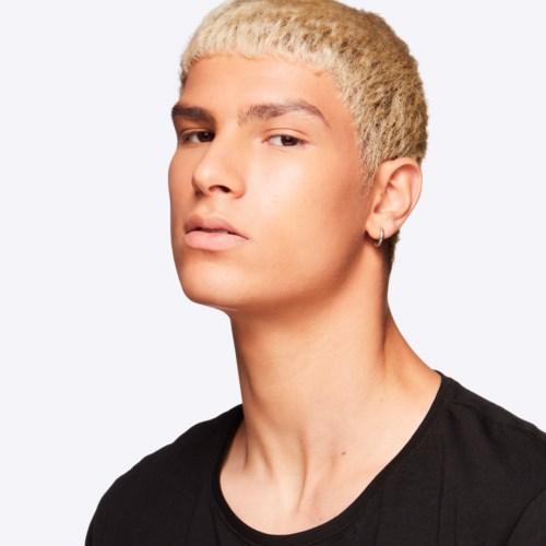 En ung person med blond hår og piercing har på sig en sort skjorte, ser alvorlig ud mod en enkel baggrund.