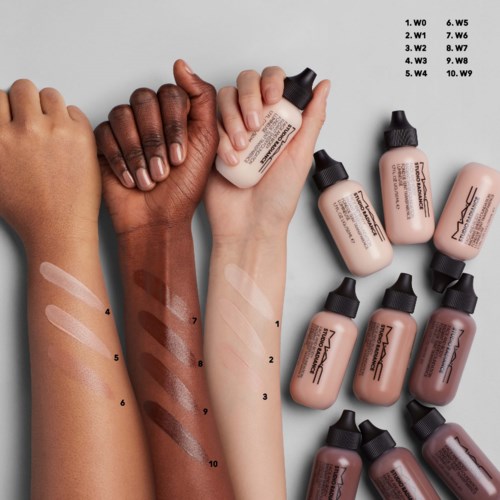 To hænder med forskellige hudtoner holder makeup foundation-flasker, nogle er swatchet på arme med tal. Flere flasker ligger i nærheden.