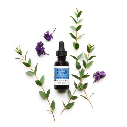 A small dark bottle labeled 'Mad Hippie' surrounded by green leaves and purple flowers.