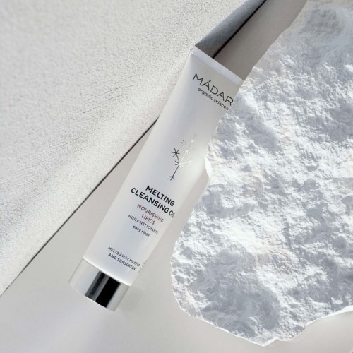 En vit tub med Melting Cleansing Oil ligger på en vit texturerad yta med en hög bomull runt den.