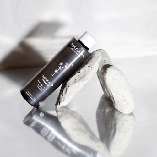 En svart flaska Madara Clarifying Toner vilar på vita stenar, kastar skugga i en ren, minimalistisk miljö.