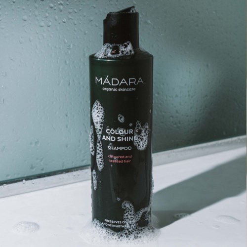 Svart flaska med MÁDARA-schampo med vita bubblor, placerad på en yta med lite skum, nära en våt spegel.