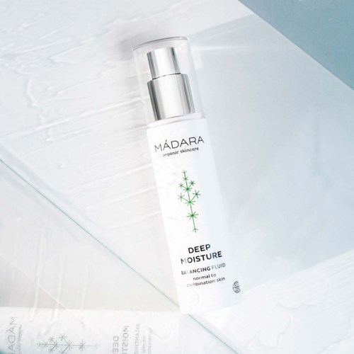 En hvit flaske med sølvpumpen, merket 'MÁDARA Deep Moisture Balancing Fluid', ligger på en hvit teksturerte overflate.