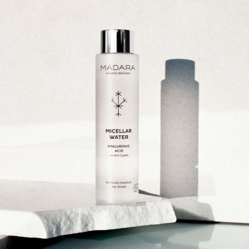 En vit flaska märkt Madara Micellar Water, med ett silverlock, står på en texturerad yta.