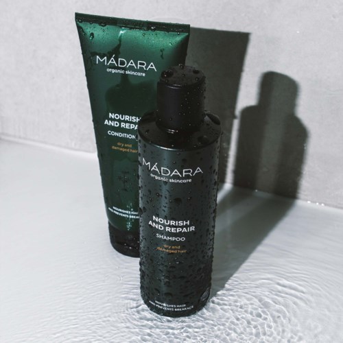 Två svarta flaskor märkta 'MÁDARA' innehåller schampo och balsam, placerade på en fuktig yta med droppar och ett skugga bakom.