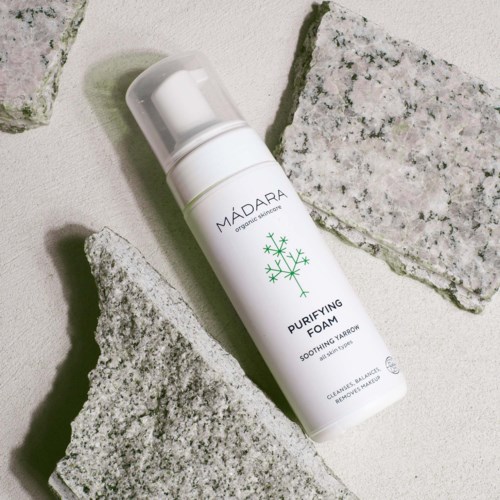 Een witte fles met 'Purifying Foam' erop, omgeven door grijze stenen op een lichte achtergrond.