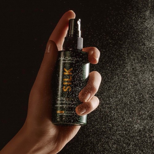 Eine Hand sprüht Nebel aus einer schwarzen Flasche mit der Aufschrift 'SILK' gegen einen dunklen Hintergrund.