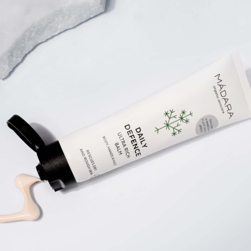 En hvit tube merket 'Daily Defence Ultra Rich Body Balm' med svart lokk, litt presset med krem som drypper ut.
