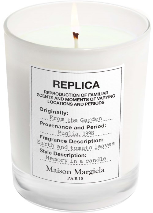 A white candle in a glass holder with a label showing scent details and brand name.