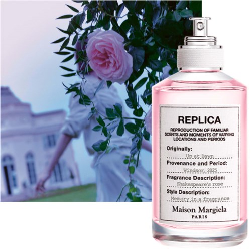 A perfume bottle labeled Replica with pink liquid, in front of a person holding a pink rose and greenery.