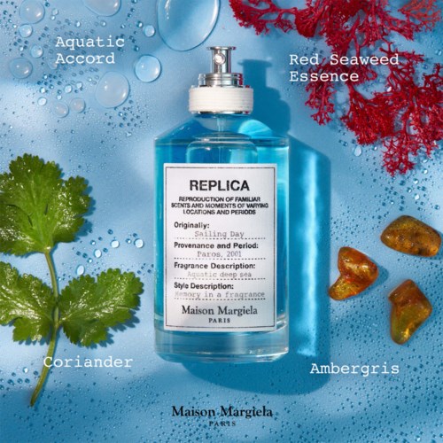 A blue perfume bottle labeled Maison Margiela Replica, surrounded by seaweed, coriander, ambergris, and water drops, on a blue background.