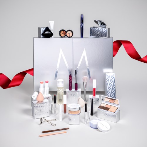 En samling av makeup-produkter, borstar och verktyg arrangerade på en vit yta med lådor och ett rött band.