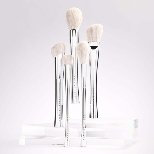 Set of five makeup brushes with white bristles and shiny silver handles, displayed on a white stand.