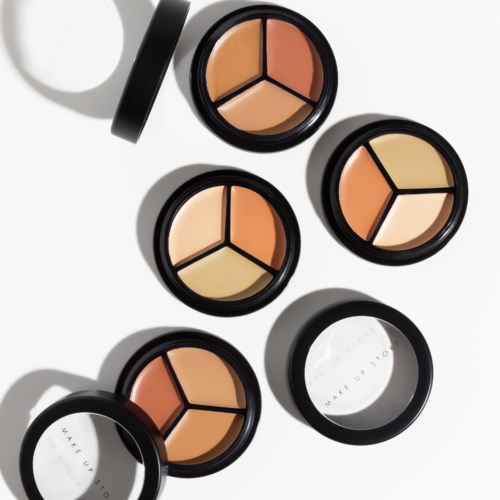 Runda makeupförpackningar med krämrouge och puder inuti, några öppna och några stängda, kastar skuggor på en vit bakgrund.