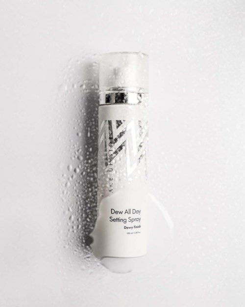 En vit sprayflaska med silverlock, märkt Dewy All Day Setting Spray, omgiven av vattondulat.