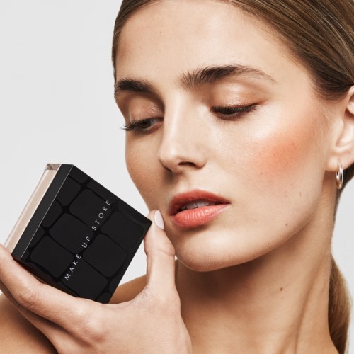 En kvinna håller i en makeupprodukt nära sitt ansikte och tittar på den med ett mjukt uttryck.