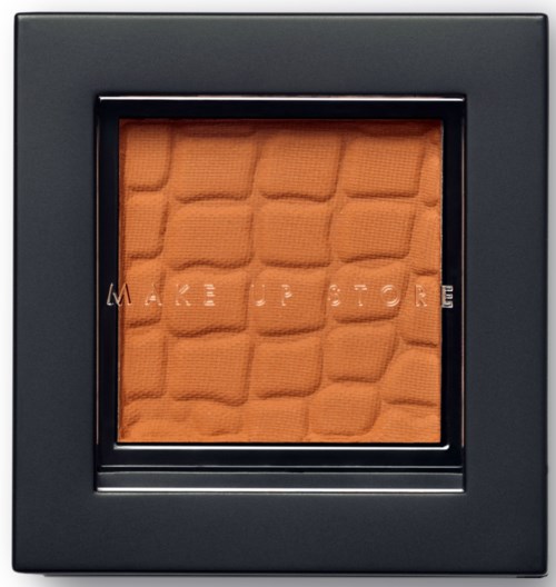 En makeup-kompakt med ett varmt orange puder inuti, omgiven av ett svart fall, och orden 'MAKE UP FOR' syns svagt.