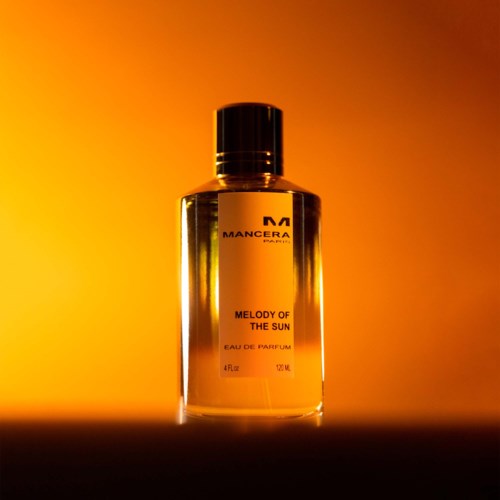 En parfahånd står mod en orange-guld gradientbaggrund, mærket "MANCERA" med duften "MELODY OF THE SUN".