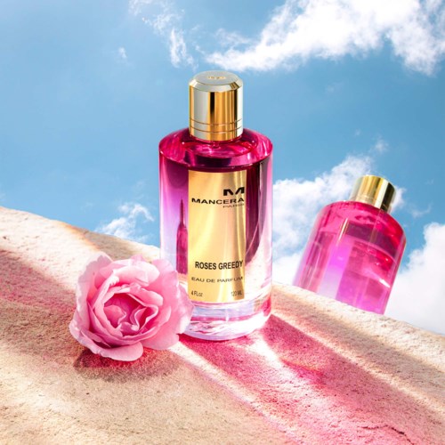 Två rosa parfymflaskor med en rosa ros på ett mjukt, rosa tyg under en blå himmel. Den ena flaskan är märkt 'MANCERA'.