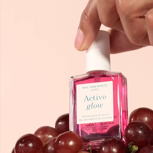 En hånd holder en rosa neglelakkflaske merket 'Active glow' blant druer.