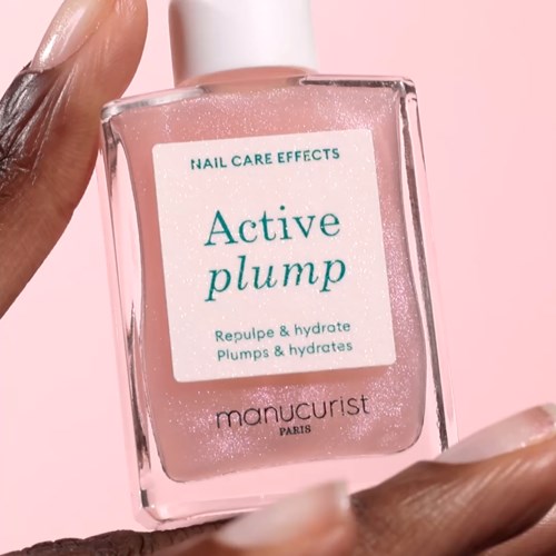 A hand holds a clear bottle of pink nail care product called 'Active plump' with a pink background.