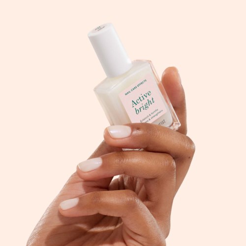 A hand holds a white bottle of nail care product called Active Bright against a light background.