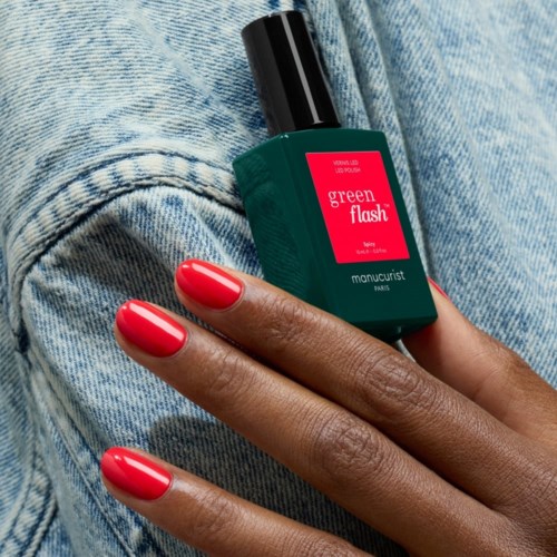 Een hand met rode nagels gelakt houdt een groene nagellakfles vast, rustend op jeans.