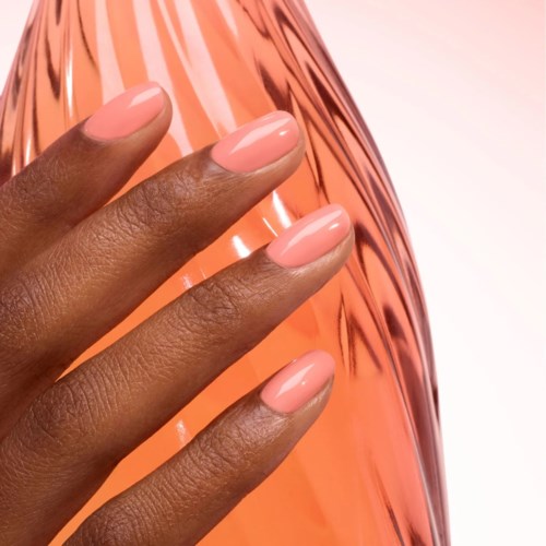 Nahaufnahme einer Hand mit gepflegten Nägeln in hellrosa Nagellack vor einem glänzenden roségoldenen Hintergrund.