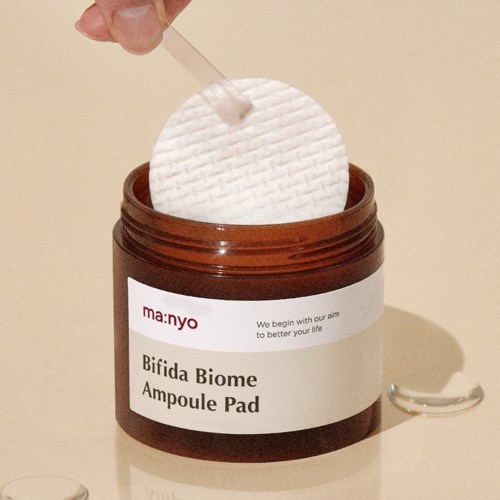 A brown jar labeled 'Bifida Biome Ampoule Pad' with a white pad being placed inside using a stick.
