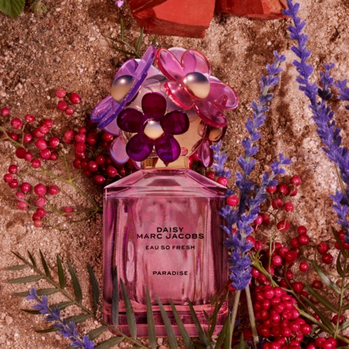 En rosa parfymflaska omgiven av lila och rosa blomformade tillbehör, röda bär och gröna blad på jord.