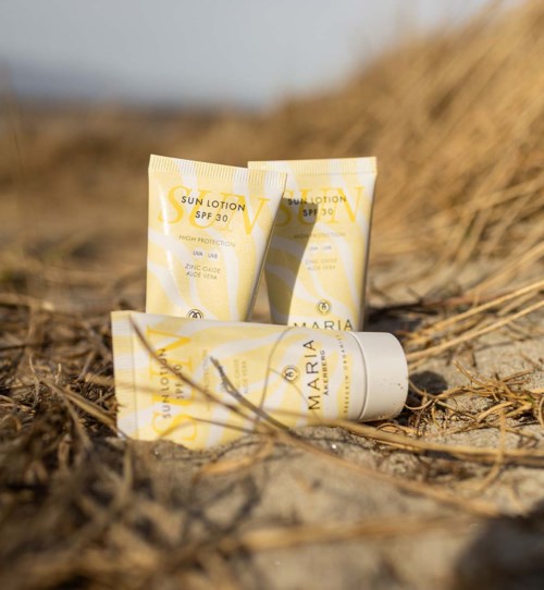 Kolme aurinkorasvatubia kuivalla niityllä, tausta hämärrän. Tubit ovat keltaisia ja valkoisia, merkattu 'Sun Lotion SPF 30' ja 'Maria'.