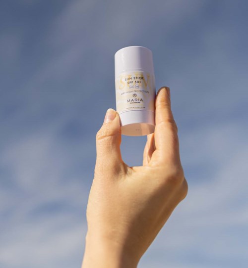 A hand holds a white sunscreen stick against a blue sky background.