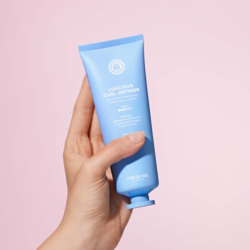 En hånd holder en blå tube merket Luscious Cure Definer av Maria Mila mot en rosa bakgrunn.