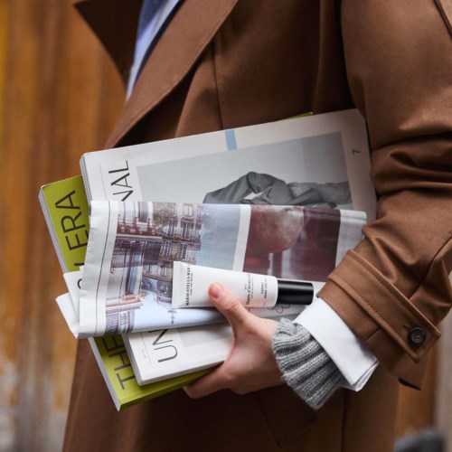 Person hält Zeitschriften und eine Tube, trägt einen braunen Mantel und grauben Ärmel. Der Hintergrund besteht aus großen Holzwänden.
