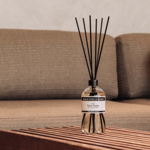 A perfume bottle with black sticks stands on a wooden surface in front of a beige couch.