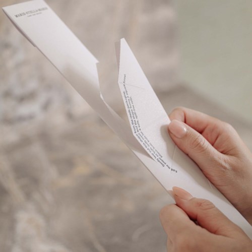 Eine Hand hält ein gefaltetes Stück Papier mit kleinem Text vor unscharfem Hintergrund.