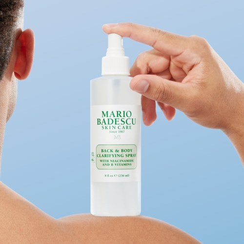 En hånd sprayer Mario Badescu hudpleiespray på en persons skulder. Flasken er hvit med grønn tekst.