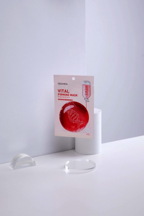 Ihonhoitotuote nimeltä 'Vital Firming Mask' punaisessa ja valkoisessa pakkauksessa, asetettuna valkoiselle pinnalle pienenä jalustana.