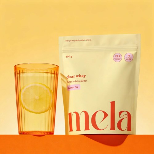 Eine weiße Tüte mit Proteinpulver Mela Zitrone neben einem Glas Zitronenwasser auf gelbem Hintergrund.