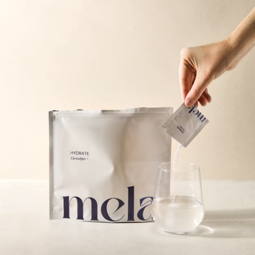 En person häller pulver från en påse i ett glas vatten. Det finns en vit påse med lila text bredvid.