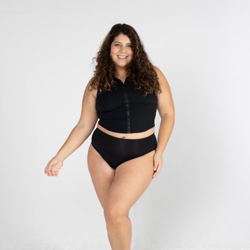 A woman with curly hair wearing a black sleeveless top and high-waisted bottoms, smiling against a white background.