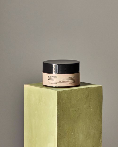 A jar of Meraki skincare sits on a green, textured block against a plain gray background.