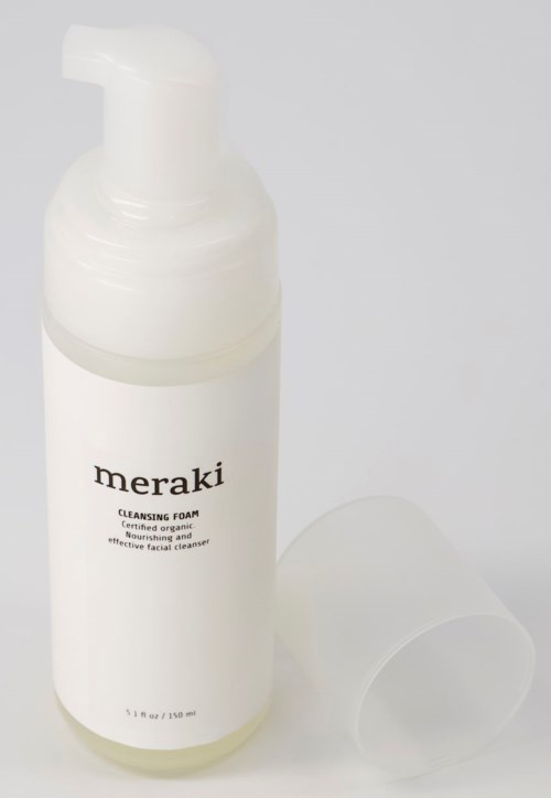 A white bottle with a pump, labeled 'meraki cleansing foam,' and a clear plastic cup beside it.