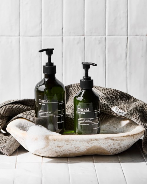 Two dark green bottles with pumps sit in a soap dish on a tiled bathroom wall. A towel is draped nearby.
