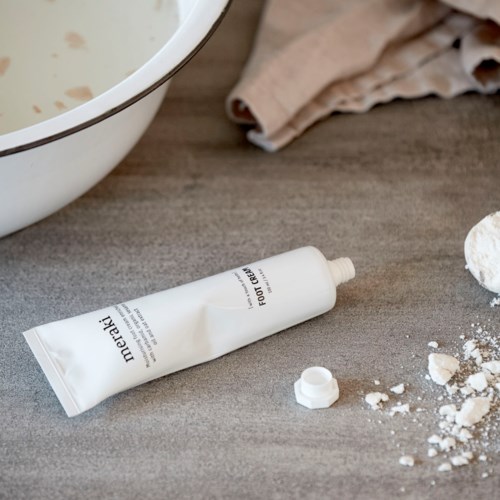 Ett vitt rör med etiketten 'Foot Cream' ligger på en trämöbel tillsammans med lite spilld puder och en handduk i närheten.