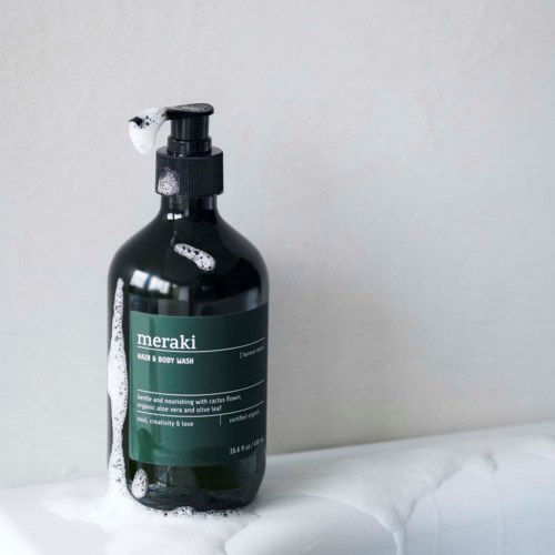 A black bottle of Meraki Hair & Body Wash with foam on it, placed on a white surface against a plain wall.