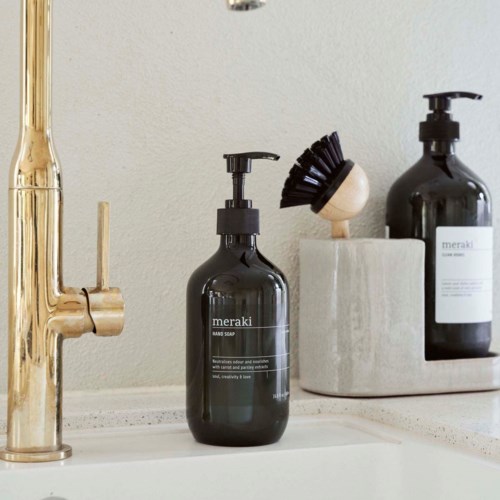 Twee zwarte flessen met 'meraki'-label staan op een witte ondergrond naast een gouden kraan. De flessen zijn voor zeep en lotion.