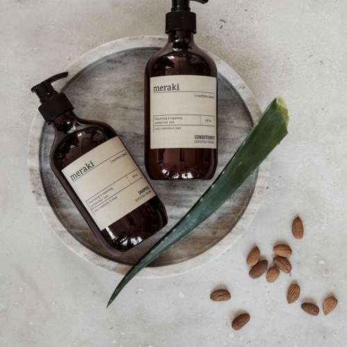 Two brown bottles of shampoo and conditioner on a round tray, with a green aloe vera leaf and some seeds nearby.