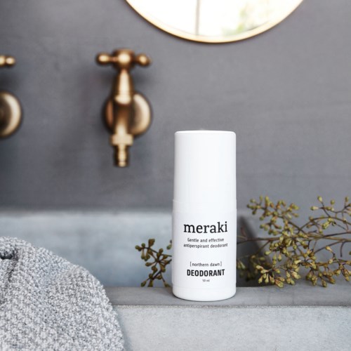 En vit Meraki deodorantflaska på ett badrumskant med spegel och guldarmaturer i bakgrunden.